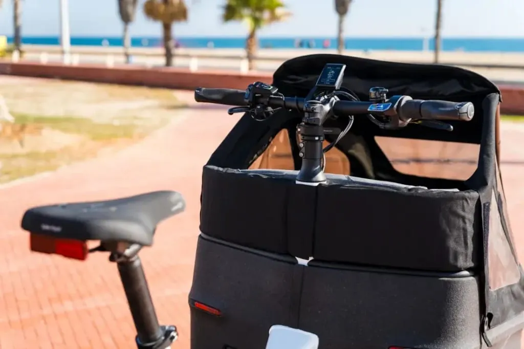 Primer plano de una bicicleta eléctrica Riese & Müller Packster2 70 CT Vario estacionada elegantemente en un sendero junto a la playa. Su manillar y asiento se destacan contra el sereno fondo del océano, ideal para familias urbanas que buscan combinar la relajación al aire libre con la comodidad moderna.