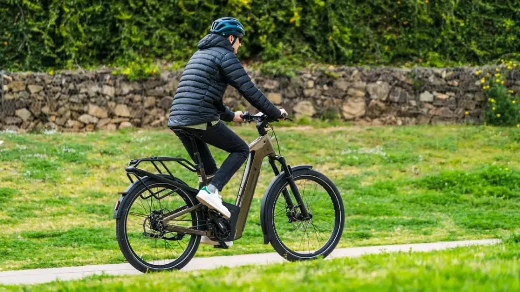 Una persona con chaqueta negra y casco conduce una bicicleta eléctrica urbana por un sendero, rodeada de vegetación y un muro de piedra. La escena encarna la tecnología de vanguardia, fusionando a la perfección la naturaleza con la innovación moderna.