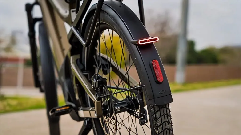 Primer plano de la rueda trasera de una bicicleta eléctrica urbana con reflector rojo y luz, estacionada en un camino pavimentado, mostrando su tecnología de vanguardia y mayor comodidad.