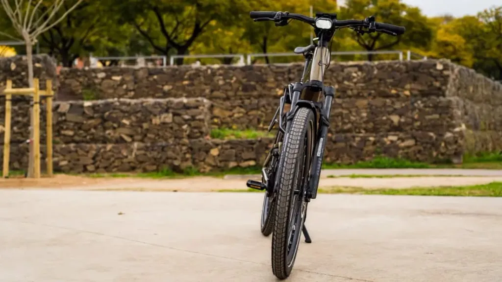 Una bicicleta eléctrica urbana descansa sobre un camino de concreto frente a un muro de piedra y frondosos árboles, combinando tecnología de vanguardia con el entorno sereno del parque.