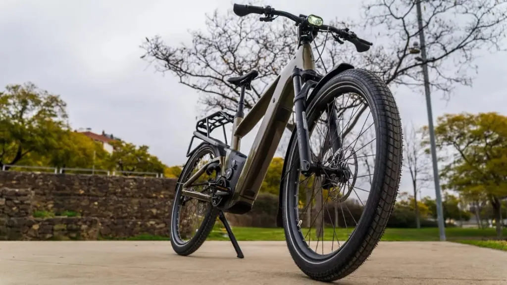 Vista de ángulo bajo de una bicicleta eléctrica urbana sobre una superficie pavimentada, enmarcada por árboles y un muro de piedra al fondo, mostrando tecnología de vanguardia en su diseño.