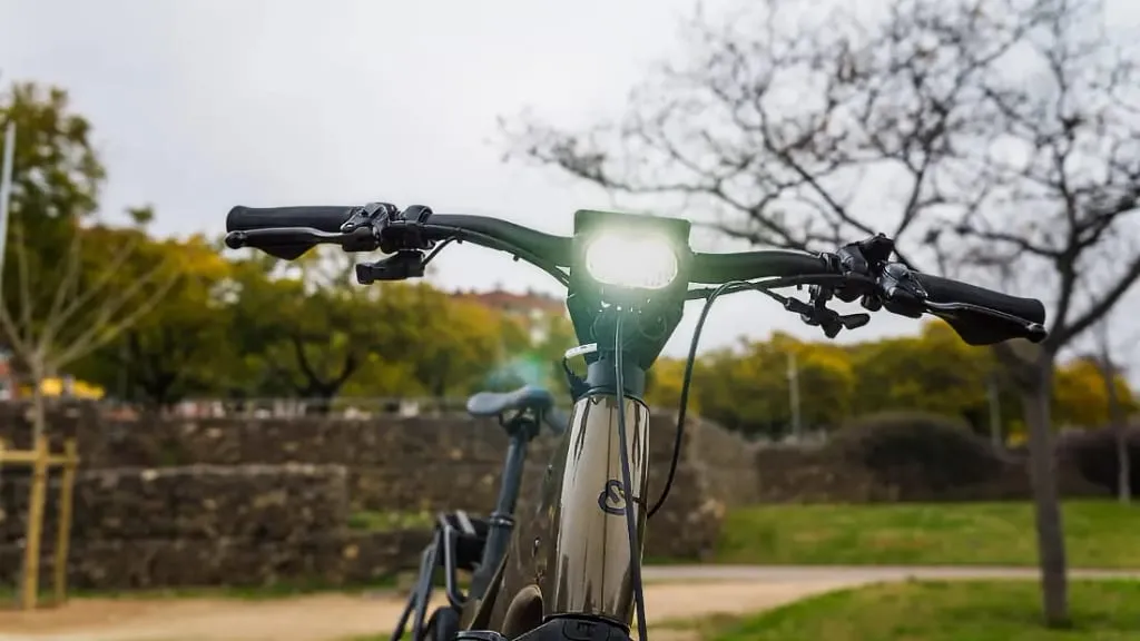 Primer plano del manillar de una bicicleta eléctrica urbana bajo una luz brillante, en un parque con árboles y un muro de piedra como telón de fondo. Disfruta de la tecnología de vanguardia mientras disfrutas de la comodidad y el estilo en tus viajes.
