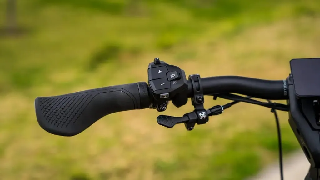 Primer plano del manillar de una bicicleta con panel de control y palancas de cambios, mostrando tecnología de vanguardia. El césped verde se ve borroso al fondo.