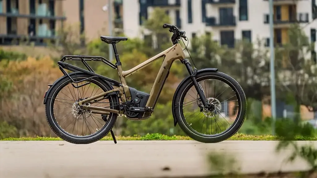 Bicicleta eléctrica urbana de color beige con portabultos trasero estacionada en un camino, ofreciendo comodidad en medio de edificios y árboles de fondo.