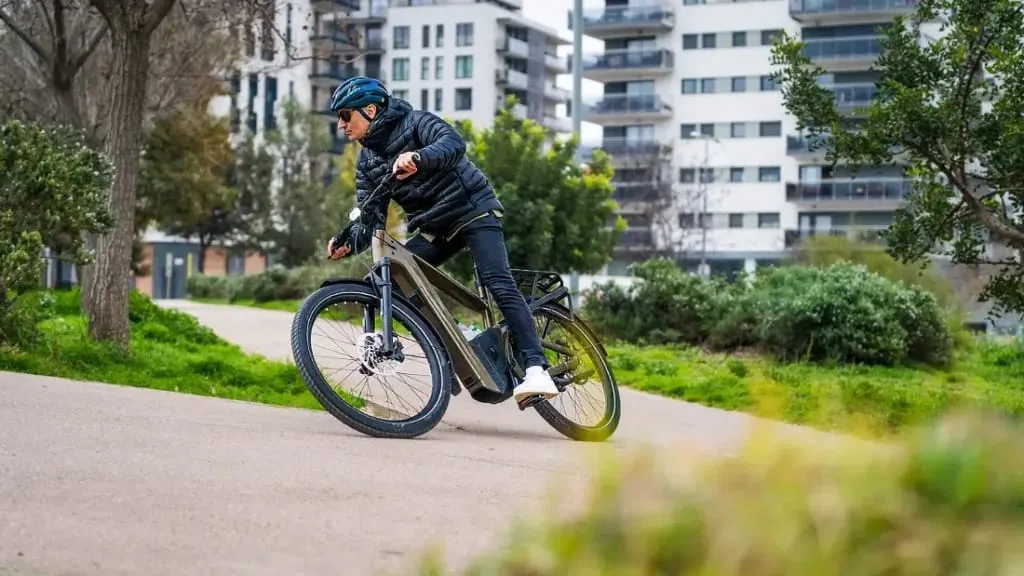 Una persona disfruta de la comodidad de una bicicleta eléctrica urbana, recorriendo el sendero de un parque con modernos edificios de apartamentos como telón de fondo, combinando a la perfección la naturaleza y tecnología de vanguardia.
