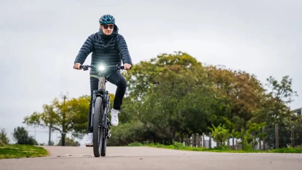 Una persona con una chaqueta negra y casco conduce una bicicleta eléctrica urbana por un sendero, envuelta por una exuberante vegetación bajo un cielo nublado.