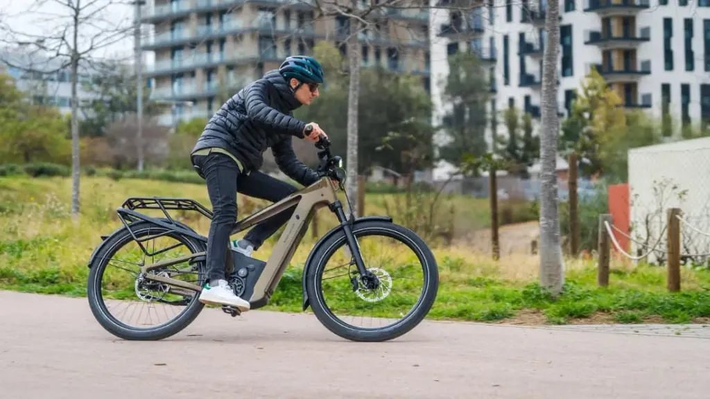 Una persona con chaqueta y casco circula por un sendero en una bicicleta eléctrica urbana, combinando a la perfección comodidad y tecnología de vanguardia, con edificios al fondo.