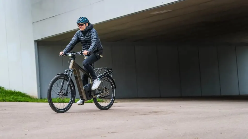 Una persona con casco y chaqueta conduce una bicicleta eléctrica, adoptando tecnología de vanguardia, en un camino pavimentado debajo de un paso elevado.