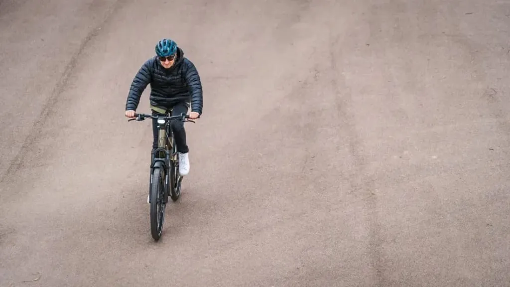 Una persona con casco y gafas de sol conduce una bicicleta eléctrica urbana por una carretera asfaltada, rodeada de un espacio abierto, disfrutando de la mezcla de comodidad y tecnología de vanguardia.