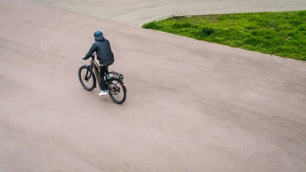 Una persona que lleva casco conduce una bicicleta eléctrica por un camino pavimentado en una zona urbana, integrándose perfectamente con el paisaje verde cercano.