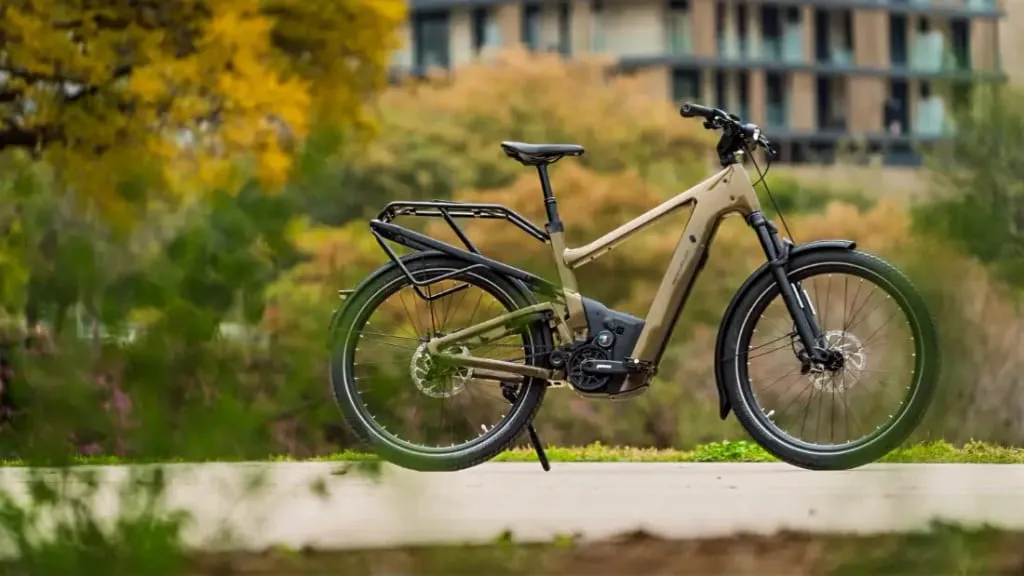 Una bicicleta eléctrica urbana de color tostado con detalles negros reposa en un camino, rodeada de árboles y un edificio al fondo, mostrando su combinación de comodidad y tecnología de vanguardia.