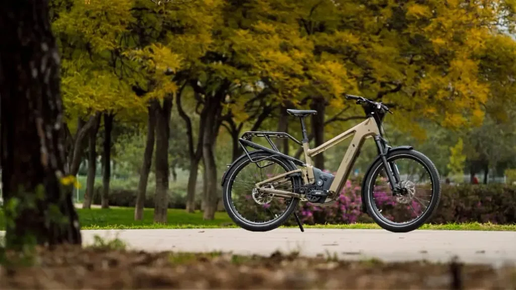 La bicicleta eléctrica urbana está estacionada en un camino, rodeada de vegetación y los tonos dorados de los árboles otoñales en un parque, combinando comodidad con tecnología de vanguardia para un paseo perfecto.
