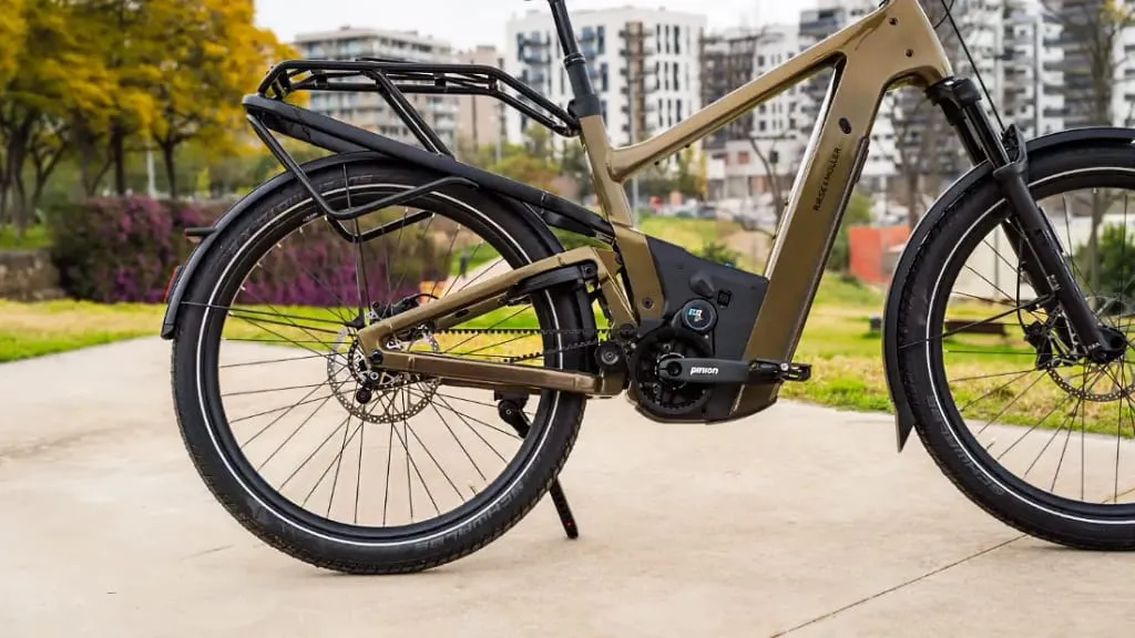 Vista lateral de una bicicleta eléctrica urbana de color beige en un camino, con un paisaje urbano y árboles al fondo, mostrando su comodidad y tecnología de vanguardia.