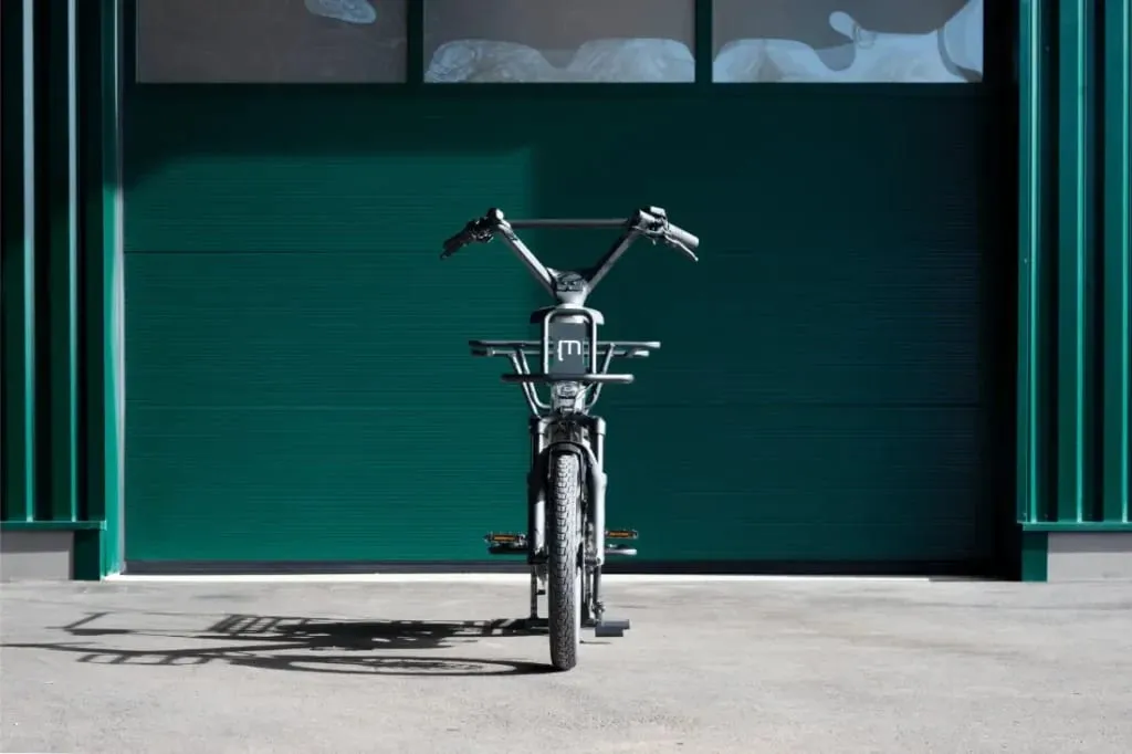 Vista frontal de una bicicleta de carga Moustache Lundi 20 con manillar alto, estacionada en la acera frente a una puerta de garaje verde.