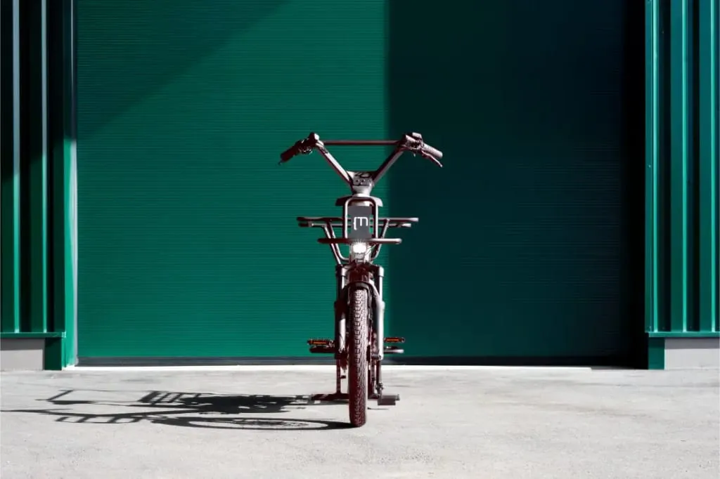 Una sola bicicleta Moustache Lundi 20 Cargo estacionada frente a una gran puerta de garaje industrial verde, vista desde el frente.