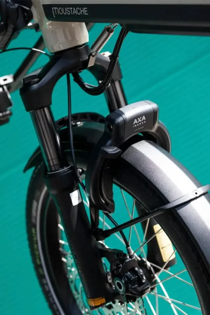 Primer plano de la rueda delantera de una bicicleta Moustache Lundi 20 Cargo con un candado AXA en la horquilla, sobre un fondo verde.