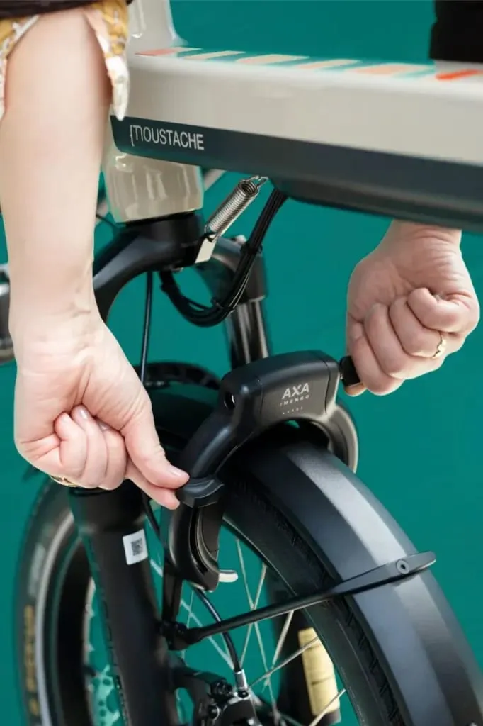 Primer plano de unas manos bloqueando la rueda delantera de una bicicleta Moustache Lundi 20 Cargo con un candado AXA.