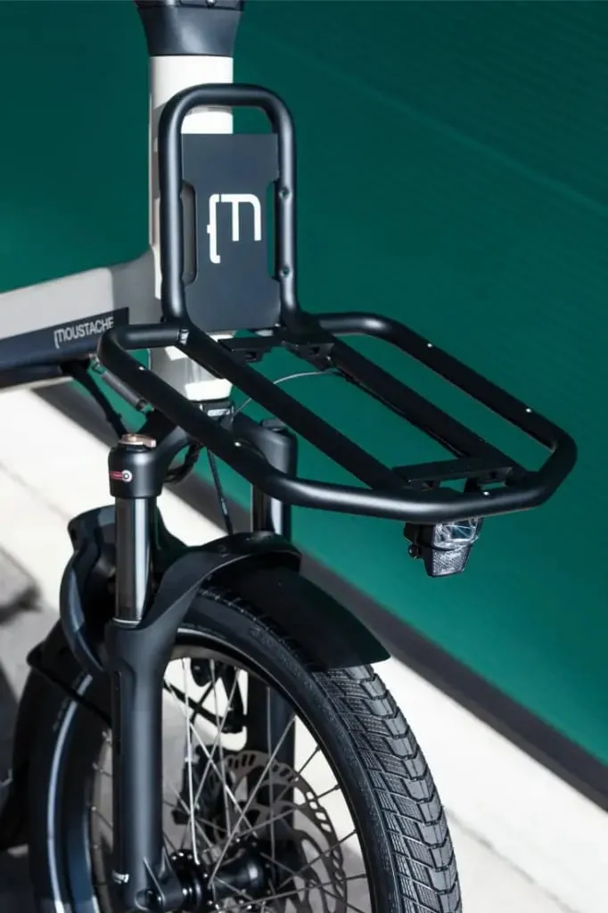 Primer plano frontal de una bicicleta de carga Moustache Lundi 20 con portaequipaje negro y faro, sobre un fondo verde.