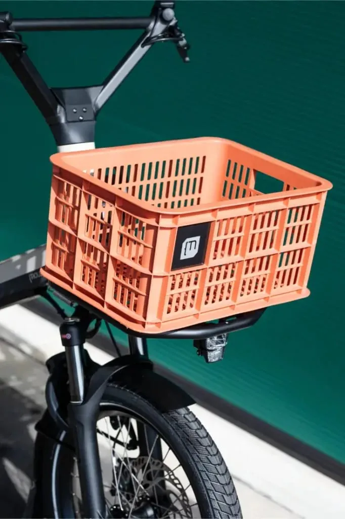 Una caja naranja está unida a la parte delantera de una bicicleta Moustache Lundi 20 Cargo negra, colocada contra una pared verde.