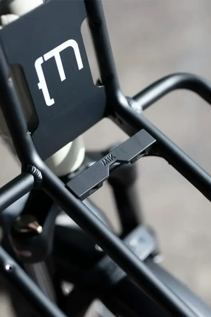 Primer plano de un portabicicletas de metal negro con un logotipo "M" y un accesorio montado, diseñado para Moustache Lundi 20 Cargo.