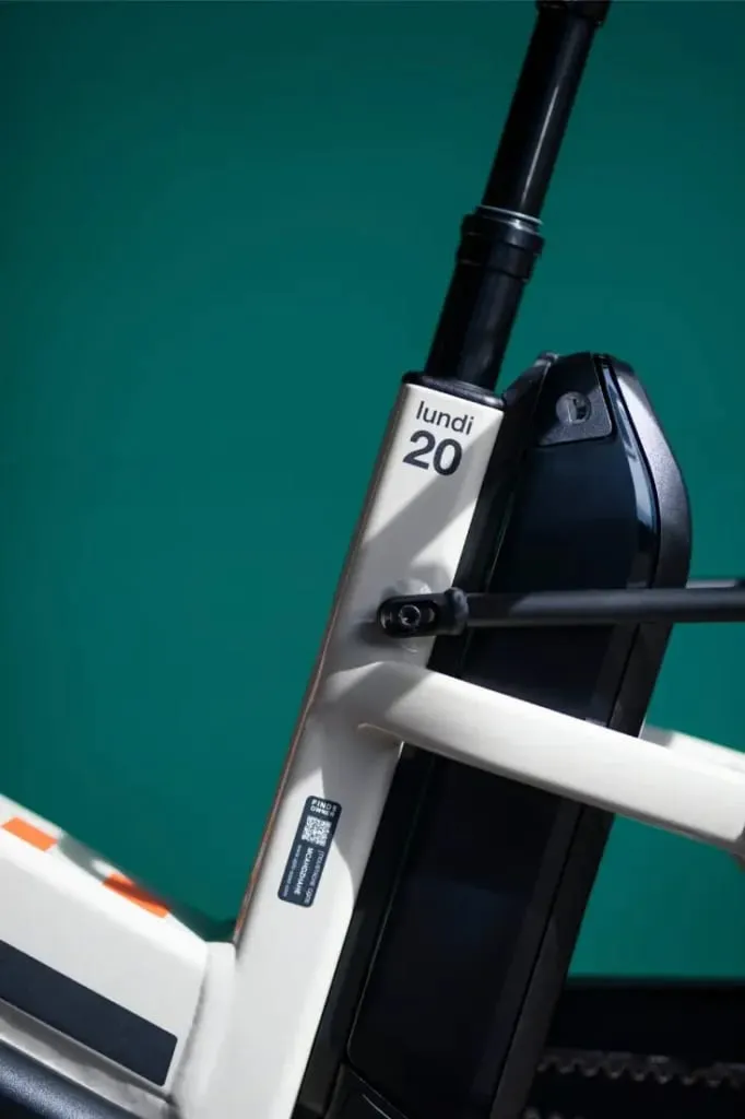 Primer plano de un cuadro de bicicleta eléctrica Moustache Lundi 20 Cargo blanco con texto "lundi 20" y una batería negra, sobre un fondo verde azulado.
