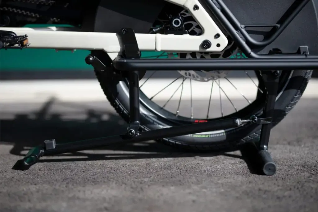 Primer plano de la rueda trasera y el caballete de un Moustache Lundi 20 Cargo, con parte del marco visible sobre una superficie pavimentada.