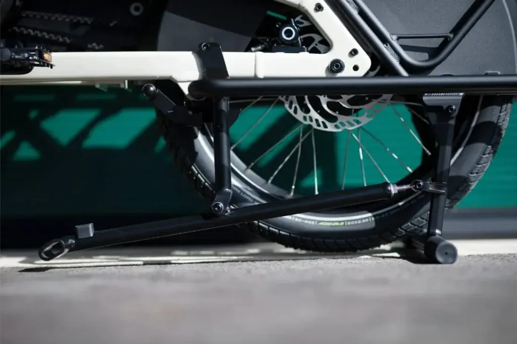 Primer plano de la rueda trasera, el soporte y el pedal de una bicicleta de carga Moustache Lundi 20 sobre el pavimento con un fondo verde.