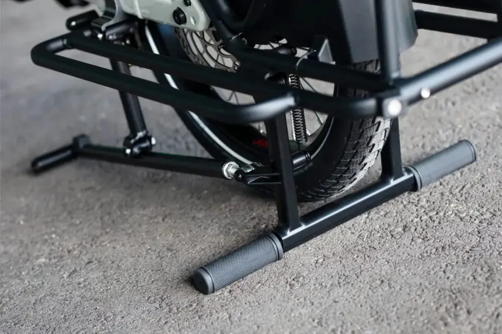 Primer plano de la rueda trasera de una motocicleta apoyada en un caballete de metal negro sobre asfalto, que recuerda al soporte resistente visto con el Moustache Lundi 20 Cargo.
