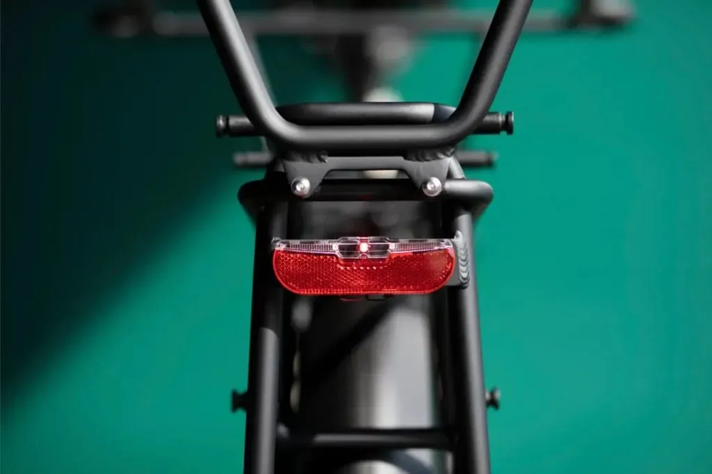 Primer plano del cuadro trasero de una bicicleta Moustache Lundi 20 Cargo con una luz trasera roja sobre un fondo verde azulado.
