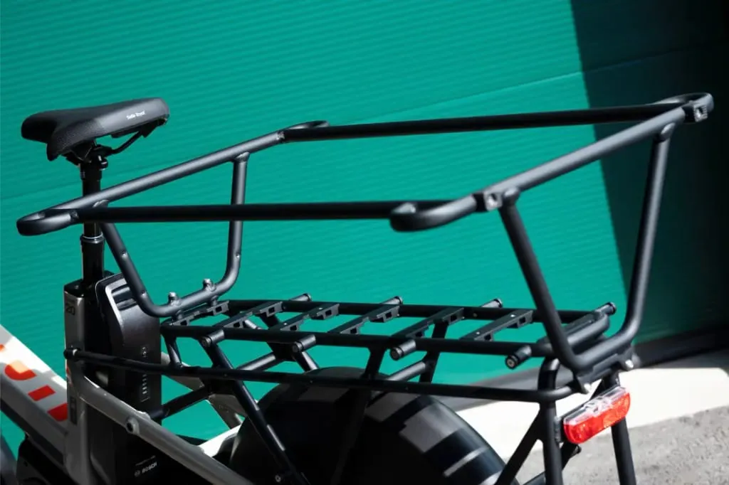 Primer plano del portaequipaje trasero negro y el cuadro de una bicicleta de carga Moustache Lundi 20 sobre un fondo corrugado verde azulado.