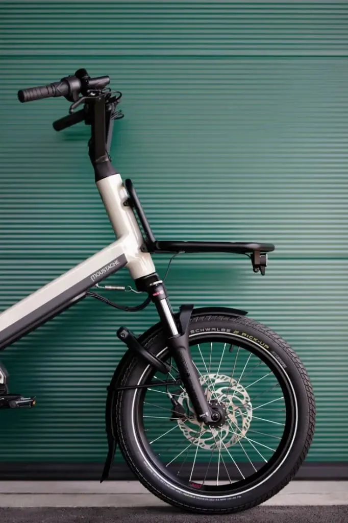 Primer plano de la rueda delantera y el manillar de una bicicleta eléctrica Moustache Lundi 20 Cargo contra una pared estriada de color verde.