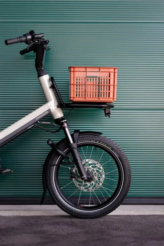 Bicicleta eléctrica de carga Moustache Lundi 20 con cesta delantera, estacionada delante de una pared metálica acanalada de color verde.