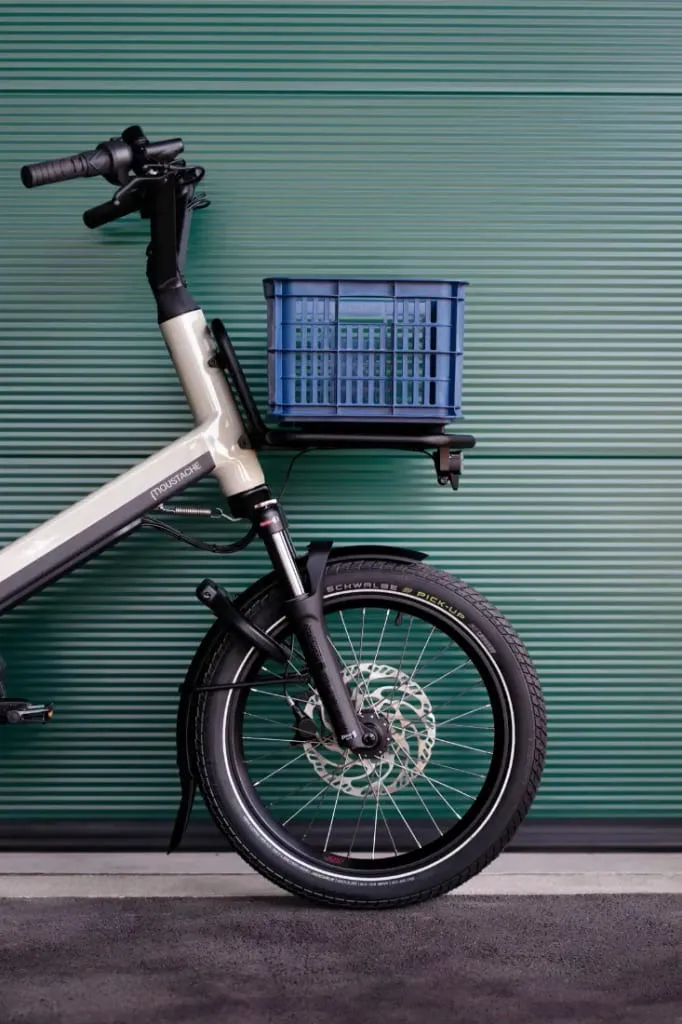 Vista frontal de una bicicleta de carga Moustache Lundi 20 con un cajón azul colocado encima de la rueda delantera, sobre un fondo verde.