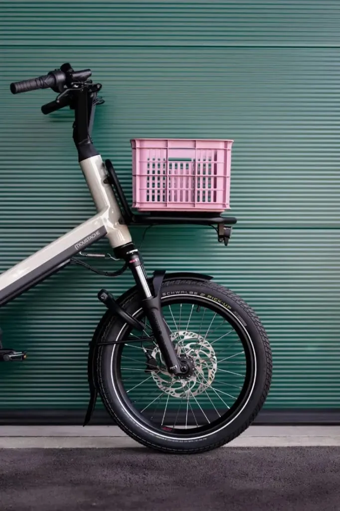 Una bicicleta de carga Moustache Lundi 20, con un cajón rosa fijado a su rueda trasera, se encuentra frente a una pared corrugada verde.