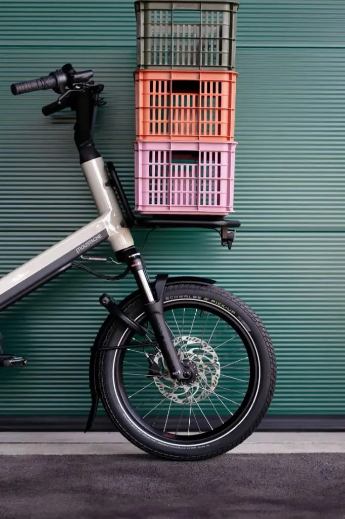 Una bicicleta de carga Moustache Lundi 20 con tres cajas apiladas montadas sobre la rueda delantera, frente a una pared de metal verde.