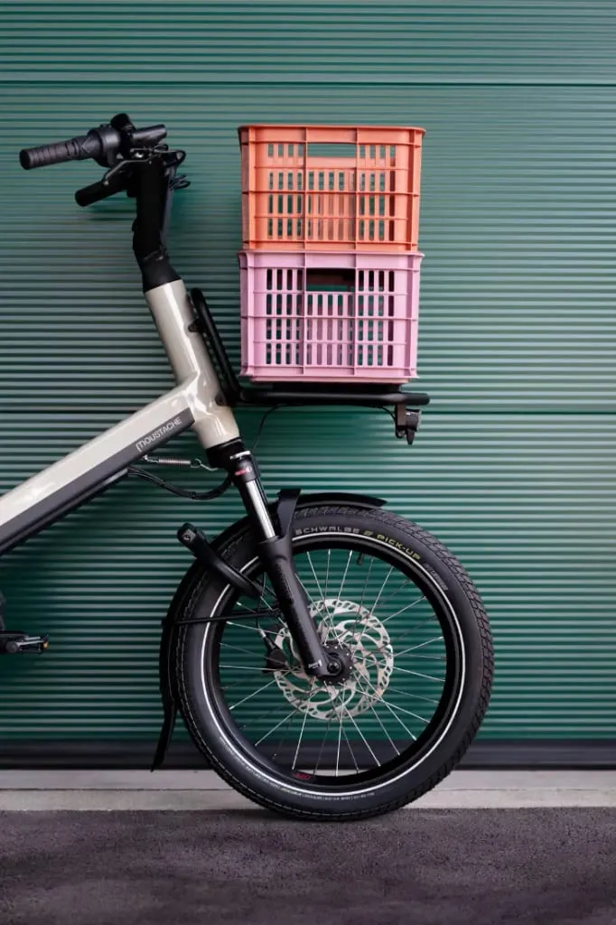 Mitad delantera de una bicicleta de carga Moustache Lundi 20 con dos cajas de plástico apiladas en un portaequipajes delantero, contra una pared verde.