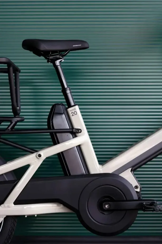 Primer plano de la bicicleta eléctrica Moustache Lundi 20 Cargo, resaltando su batería, asiento y cuadro contra una pared verde estriada.