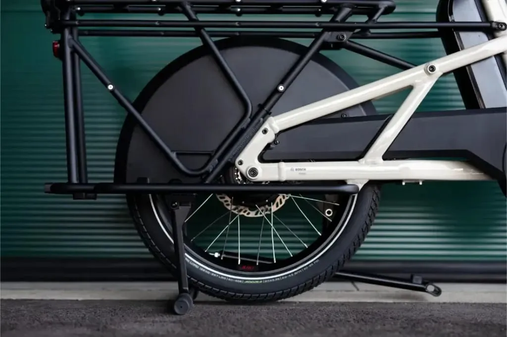 Primer plano de la rueda trasera y el cuadro de una bicicleta eléctrica Moustache Lundi 20 Cargo sobre un fondo de metal corrugado verde.