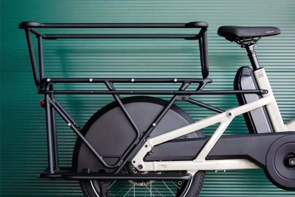La bicicleta eléctrica Moustache Lundi 20 Cargo, en color blanco, cuenta con un portaequipajes trasero de gran tamaño y está diseñada sobre un llamativo fondo corrugado de color verde.