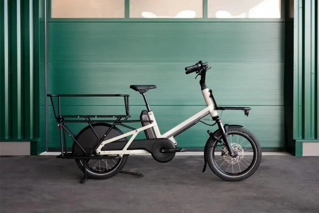 Bicicleta eléctrica de carga Moustache Lundi 20 con portabultos trasero largo, estacionada frente a una pared con paneles verdes.