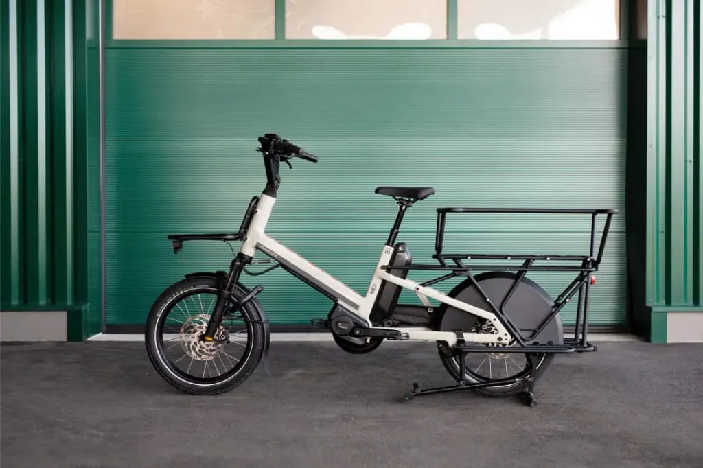 La Moustache Lundi 20 Cargo, una bicicleta de carga eléctrica blanca con portaequipaje trasero, está estacionada frente a una pared de metal verde.