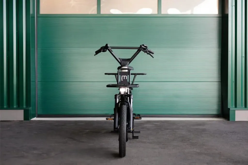 Una bicicleta eléctrica Moustache Lundi 20 Cargo negra mira hacia adelante, estacionada frente a una gran puerta de garaje de metal verde.
