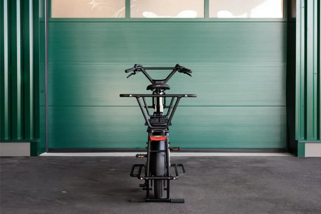 Una bicicleta de carga Moustache Lundi 20 de color negro está estacionada frente a una puerta de garaje verde cerrada sobre una superficie pavimentada.