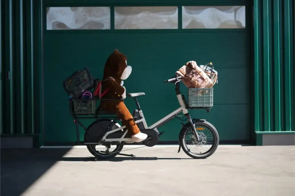 Un mono de peluche y bolsas están cargadas en una bicicleta eléctrica Moustache Lundi 20 Cargo estacionada frente a una puerta de garaje verde.