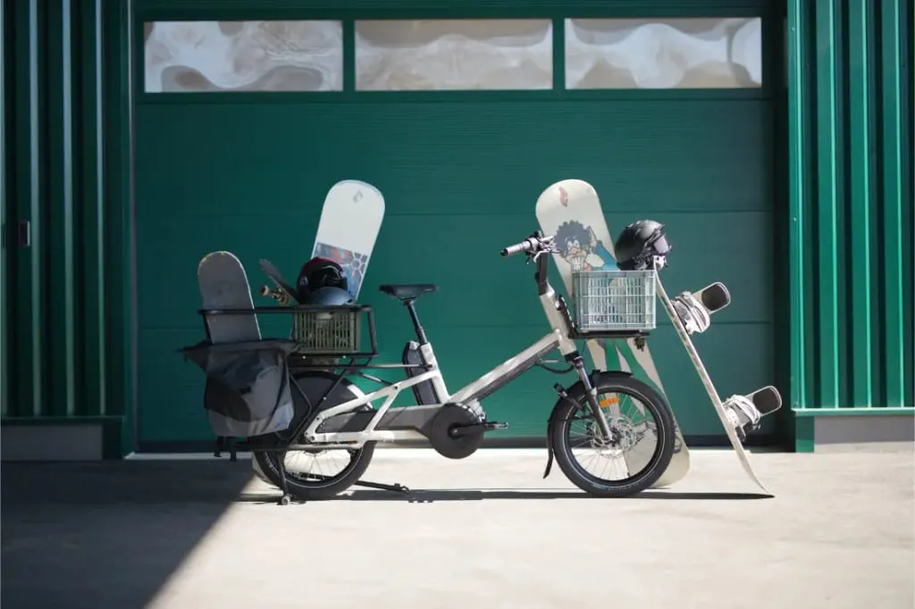 Una bicicleta de carga Moustache Lundi 20 está estacionada frente a un garaje verde, cargada con tablas de snowboard, cestas y cascos.
