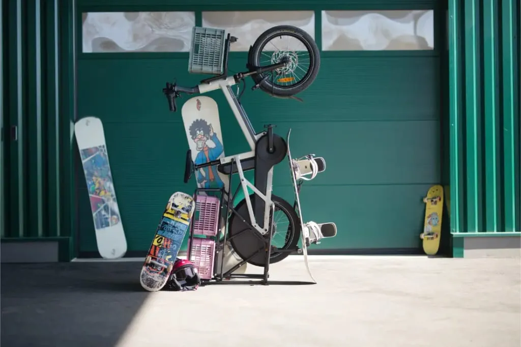 Una bicicleta de carga blanca Moustache Lundi 20 se encuentra en posición vertical sobre su manillar, rodeada de patinetas y un casco frente a un garaje verde.