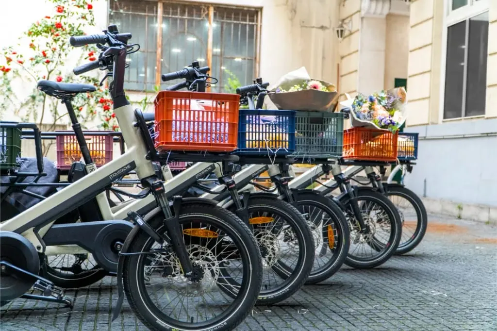 Cuatro bicicletas, incluida una Moustache Lundi 20 Cargo con una cesta delantera llena de flores, están alineadas en una calle adoquinada cerca de un edificio.