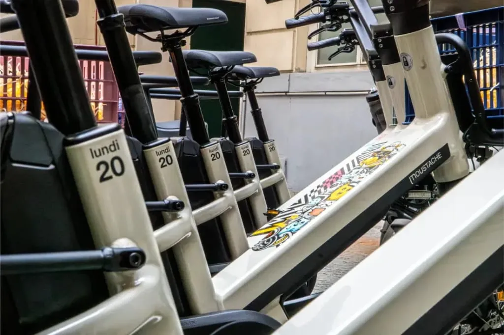Fila de bicicletas eléctricas Moustache Lundi 20 Cargo blancas, etiquetadas como "lundi 20", estacionadas muy cerca unas de otras en una calle de la ciudad.