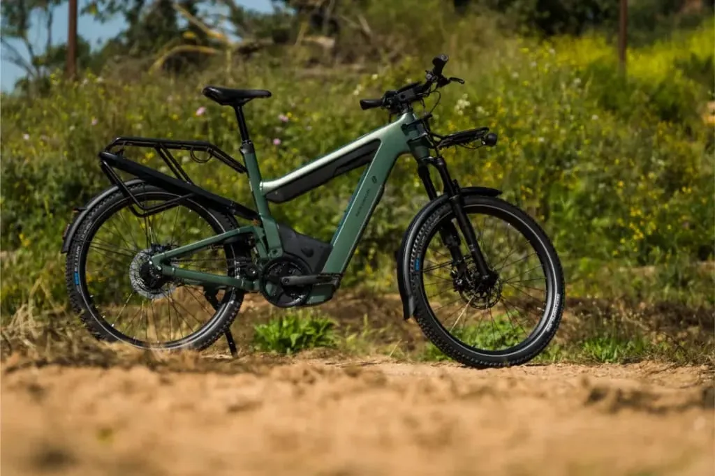 Bicicleta eléctrica verde, Riese & Müller Superdelite5, con portaequipaje trasero, estacionada en un camino de tierra rodeado de exuberante vegetación al fondo.