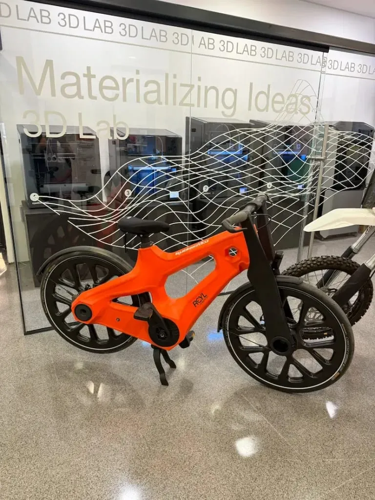 Una bicicleta igus de color naranja brillante impresa en 3D a partir de redes de pesca se exhibe en un laboratorio moderno denominado "Materializing Ideas 3D Lab", que destaca la innovación en movilidad urbana.